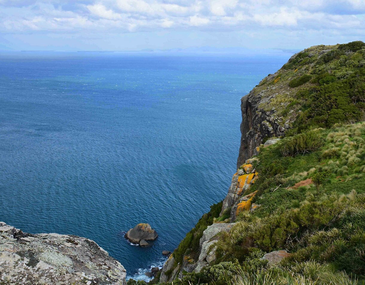 Steep green coastal cliff overlooking deep blue ocean water with orange lichen-covered rocks and a small rock formation visible in the water below.