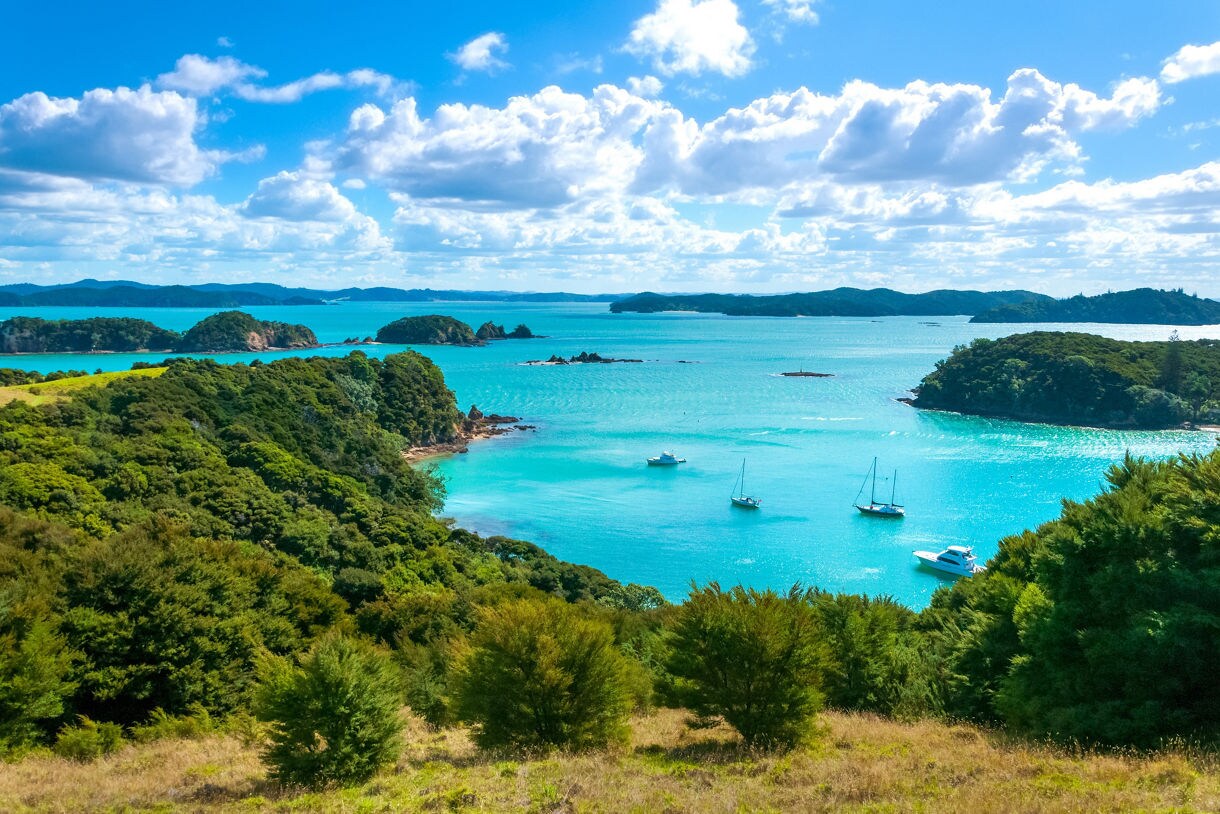 Aerial view of sailboats anchored among lush green islands in New Zealand’s Bay of Islands, surrounded by vivid turquoise water under a bright sky.
