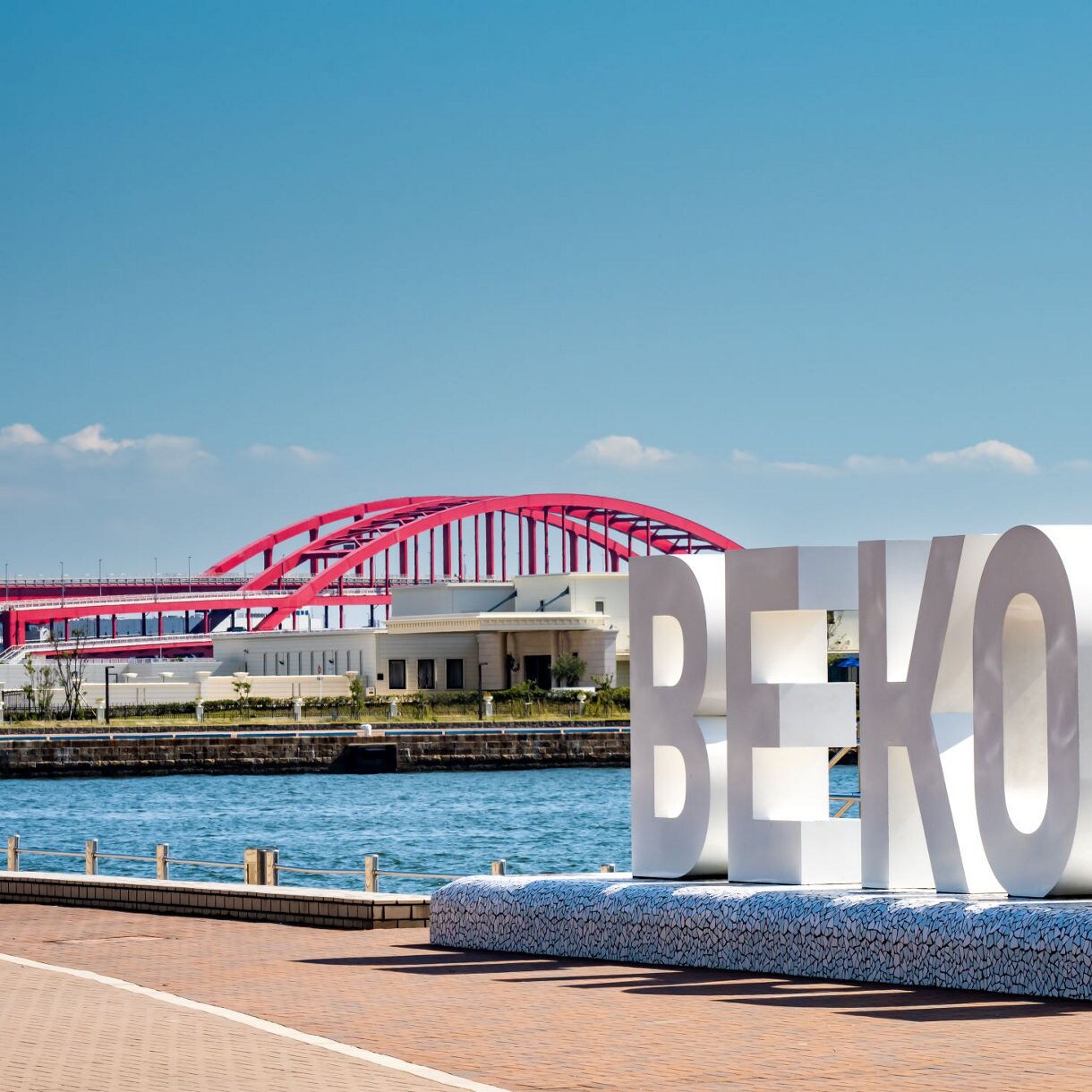 Large white 'BE KOBE' sign on a waterfront promenade in Kobe, Japan, with a red arched bridge spanning the water in the background, and city buildings under a bright blue sky with scattered clouds.