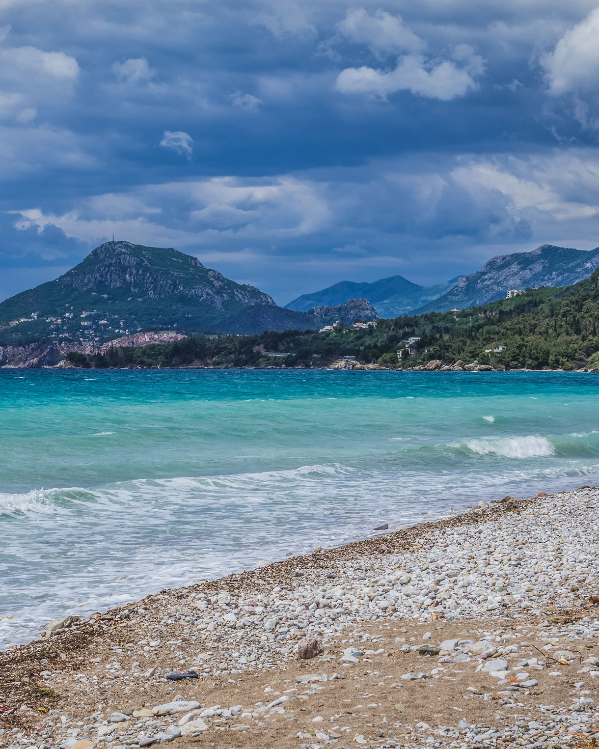 Pebble beach with gentle waves and turquoise water, backed by forested mountains under a cloudy sky in Bar, Montenegro.