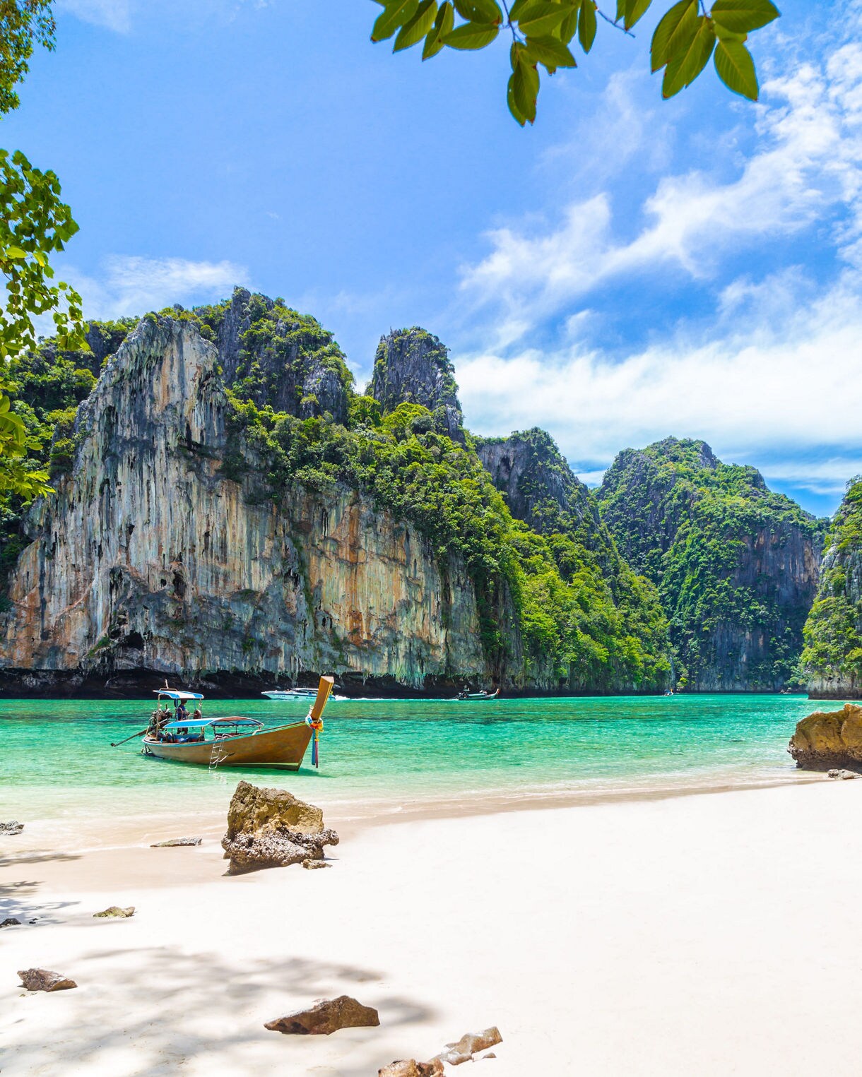 A longtail boat floating near a white sandy beach with turquoise water and towering limestone cliffs in Thailand.