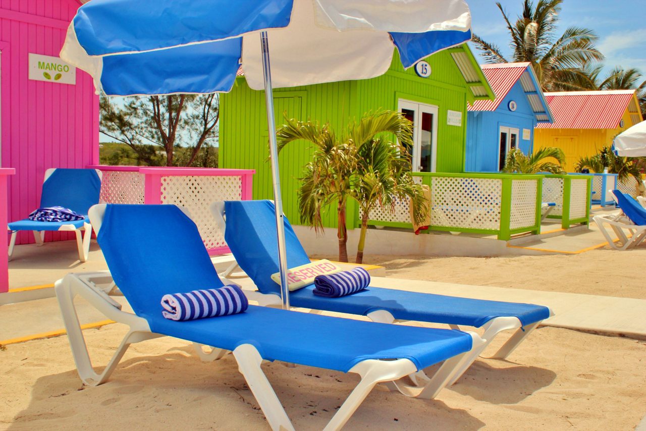 Blue beach loungers with umbrellas positioned in front of colorful pink, green, and blue beach cabanas with palm trees at Princess Cays.