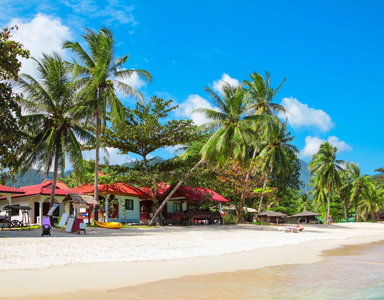 Peaceful tropical beach in Thailand lined with palm trees and small red-roofed bungalows beside calm turquoise water.