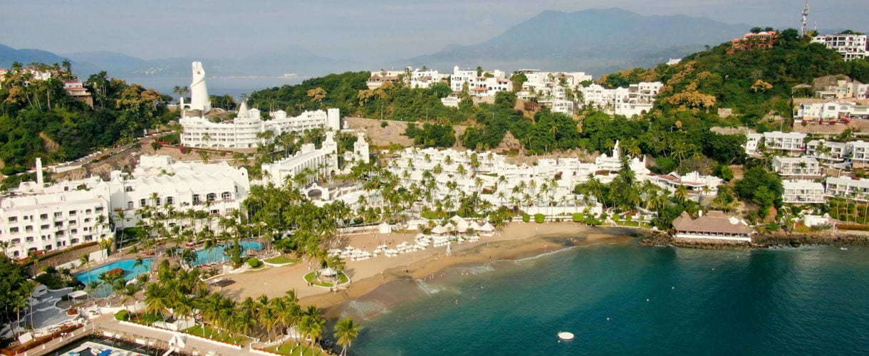 Aerial view of a beachfront resort in Manzanillo, Mexico, with white buildings, palm trees, a marina, and a sandy beach, surrounded by green hills and blue waters.