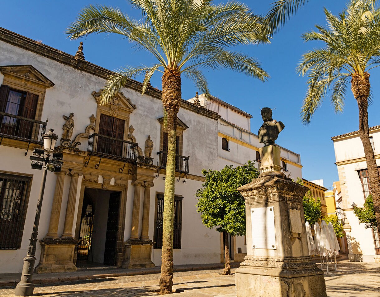 Small plaza with tall palm trees, a stone bust on a pedestal and whitewashed historic buildings with balconies under a clear blue sky.