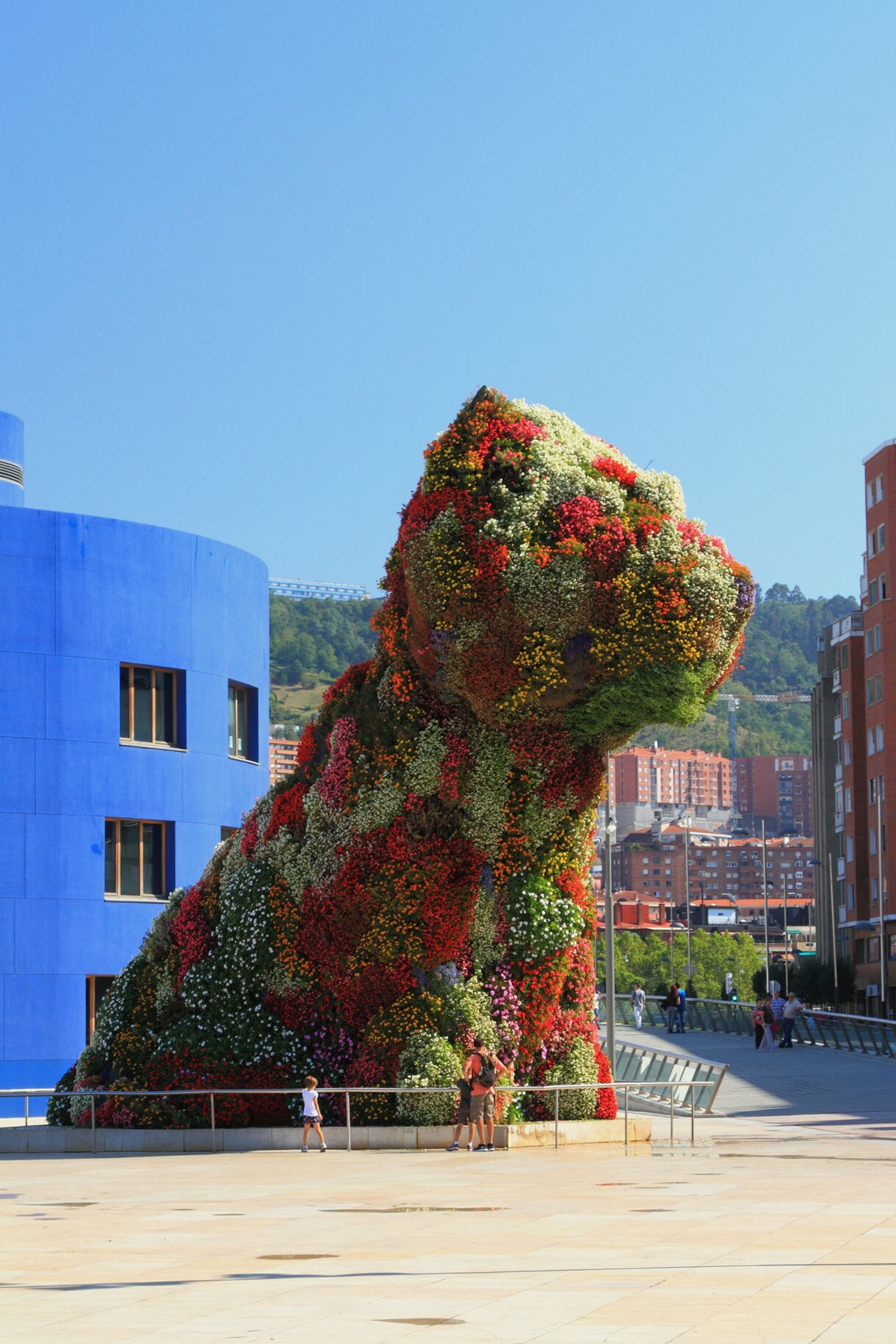 Giant dog-shaped sculpture covered in colorful flowers standing near the Guggenheim Museum in Bilbao, Spain, with people walking around its base.