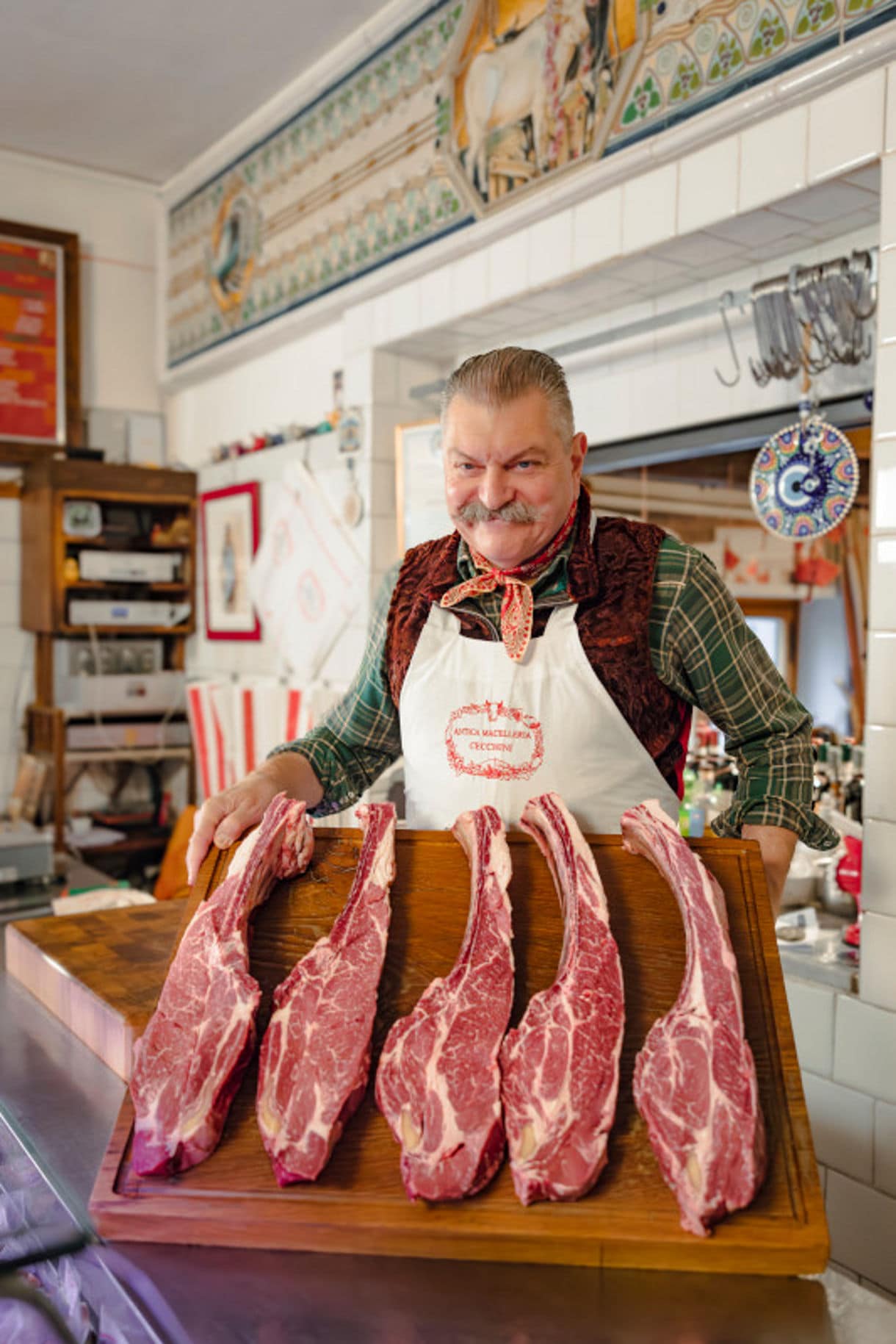 A butcher in a white apron displaying several large cuts of aged beef on a wooden cutting board.