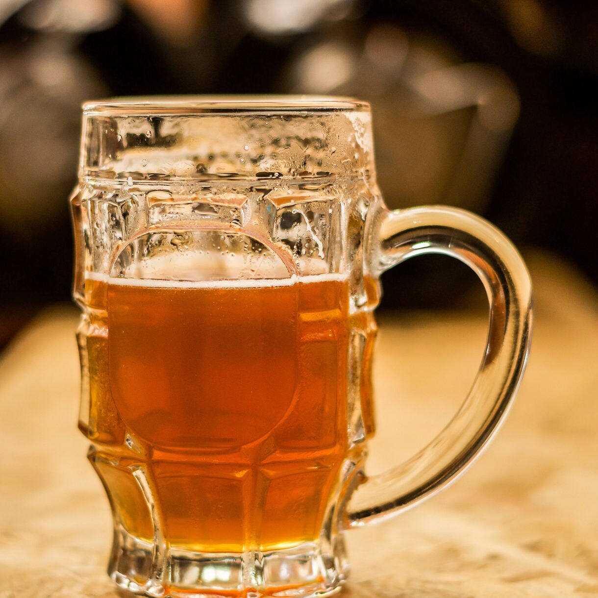 Close-up of a glass mug filled with amber beer, condensation on the sides, resting on a wooden surface with a blurred background.