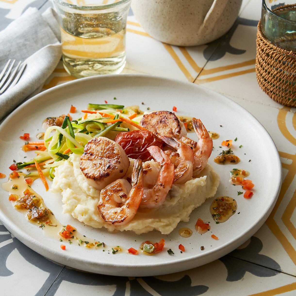A plated seafood dish featuring shrimp and scallops served over creamy mashed potatoes. The meal is garnished with colorful vegetables and herbs, presented on a patterned table setting. A glass of white wine and a folded napkin complete the elegant dining scene.