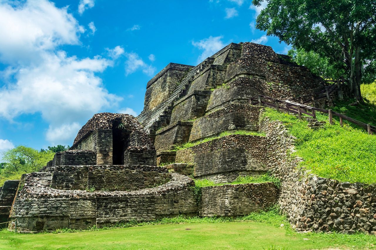 The stone ruins of Altun Ha in Belize, featuring a stepped pyramid with weathered stone walls, patches of green grass, and a wooden staircase leading to the top, set against a bright blue sky with scattered clouds.