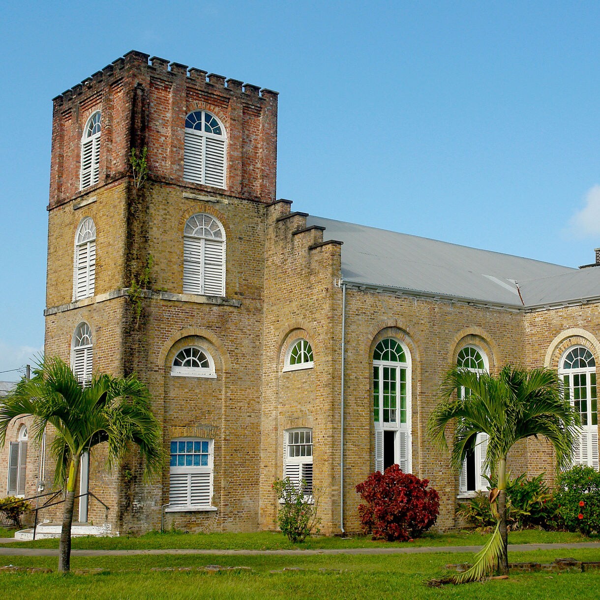 The historic Anglican Church in Belize, featuring a tall brick tower, arched windows, and surrounded by palm trees and vibrant flowers under a blue sky.