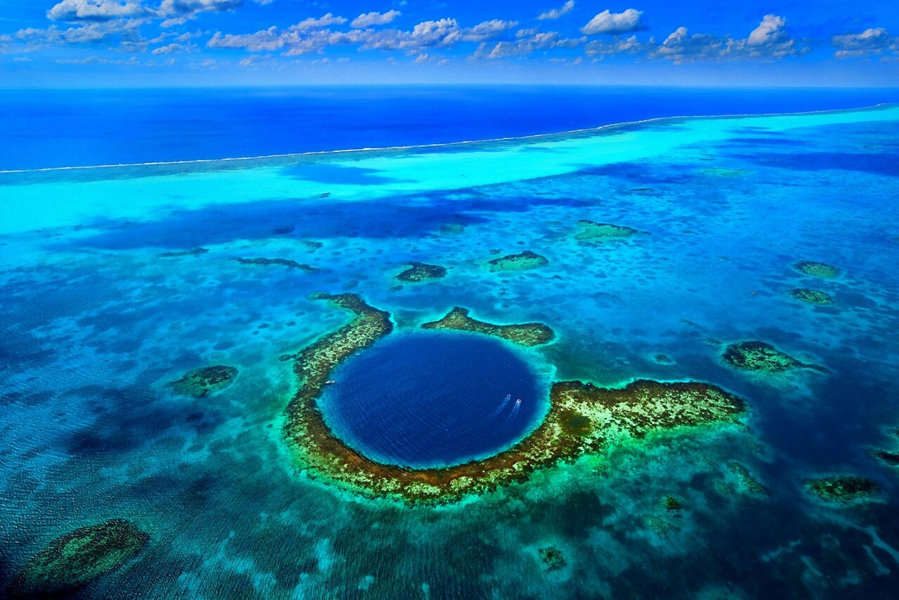 Aerial view of the Great Blue Hole, a large circular marine sinkhole surrounded by coral reefs and turquoise waters, with two boats approaching.