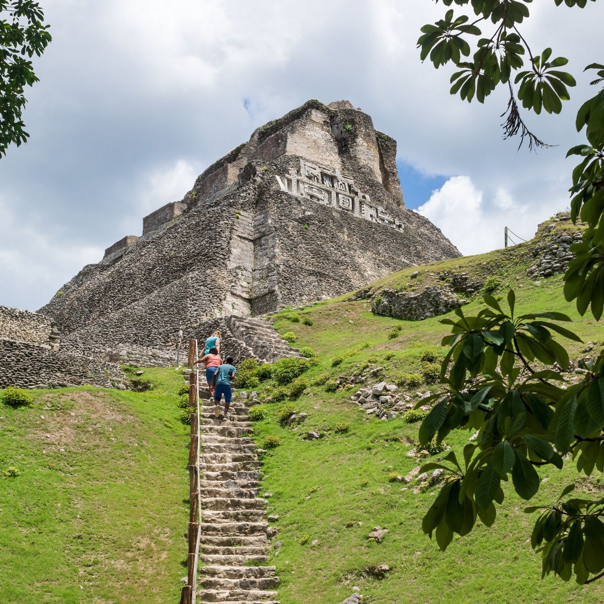 Tourists climb stone steps leading to the top of the Xunantunich pyramid, with the ancient Mayan carvings visible at the summit against a clear sky.