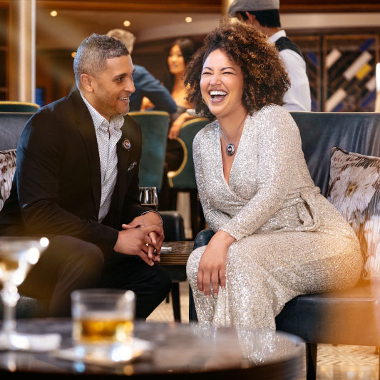 Two guests share a laugh while seated on a navy blue couch in an elegant lounge setting, with the woman wearing a sparkly silver dress and the man in a dark suit with a patterned shirt.