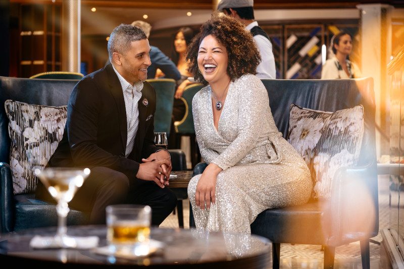 Two guests share a laugh while seated on a navy blue couch in an elegant lounge setting, with the woman wearing a sparkly silver dress and the man in a dark suit with a patterned shirt. 