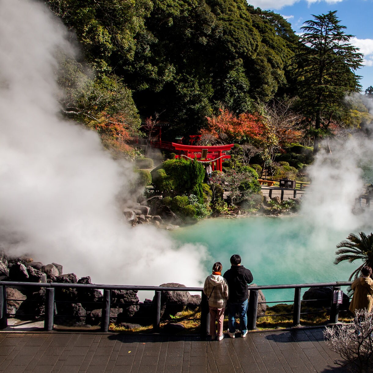 Visitors look over a steaming turquoise hot spring in Beppu, Japan, with a red torii gate and autumn foliage in the background.