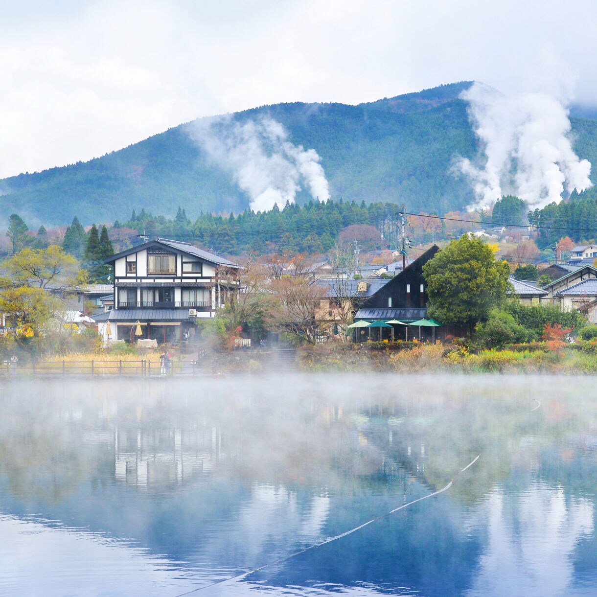 Steam rises from a calm lake in Yufuin Onsen, Japan, with traditional houses and forested hills in the background.
