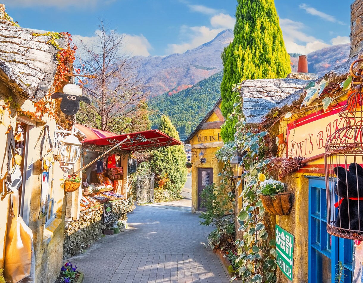 Colorful cottage-style shops line a narrow lane in Yufuin Floral Village, with mountain peaks rising in the background.
