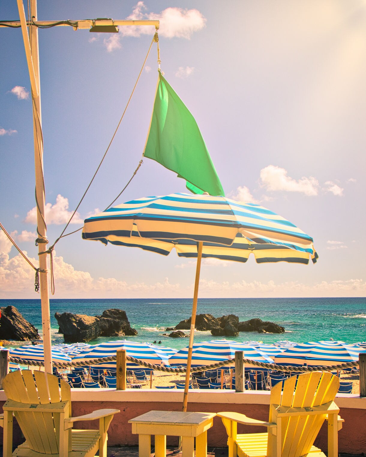 Yellow chairs under a striped umbrella overlooking the turquoise ocean and rows of beach umbrellas in Bermuda.