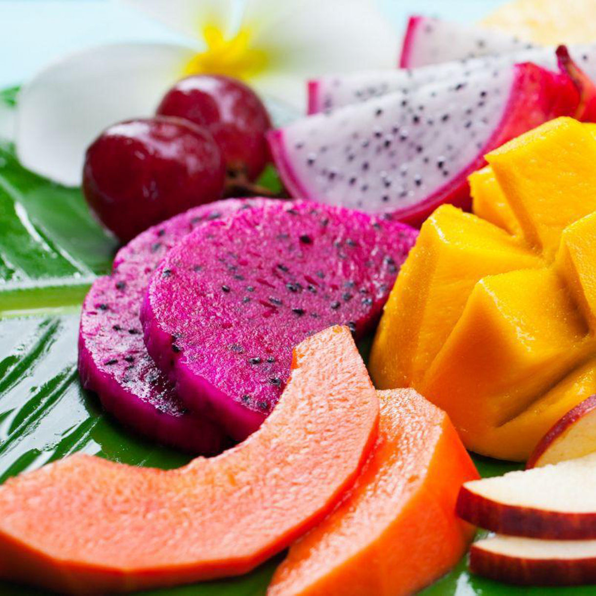 Colorful fruit platter featuring sliced mango, dragon fruit, watermelon, papaya, apple, pineapple, grapes and lychee, arranged on a large green banana leaf.