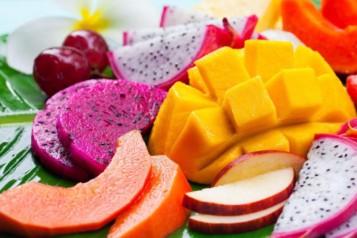 Colorful fruit platter featuring sliced mango, dragon fruit, watermelon, papaya, apple, pineapple, grapes and lychee, arranged on a large green banana leaf.