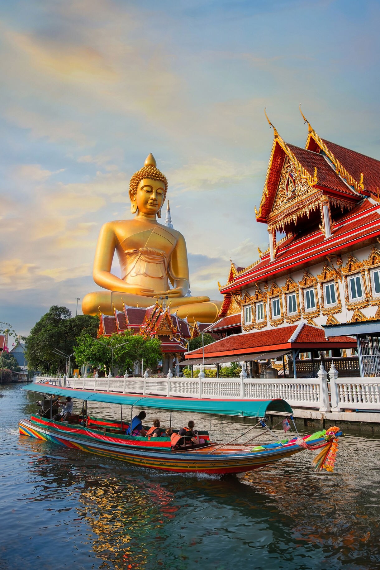 A golden seated Buddha statue overlooking a canal in Bangkok with a colorful longtail boat passing in front of a red-roofed temple.