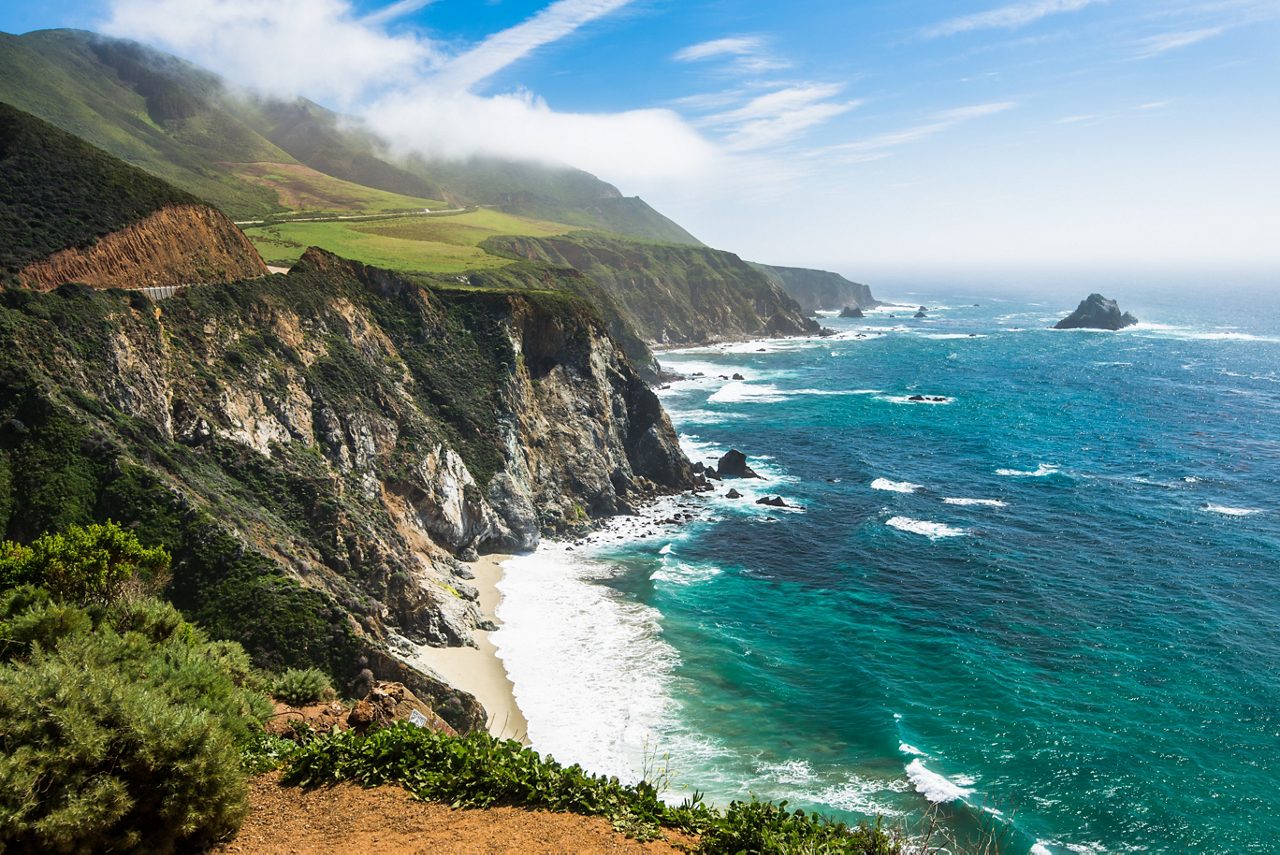 A breathtaking view of the rugged Big Sur coastline in California, with steep cliffs dropping down to turquoise waters and white sandy beaches, under a bright blue sky with patches of clouds hugging the green hillsides.