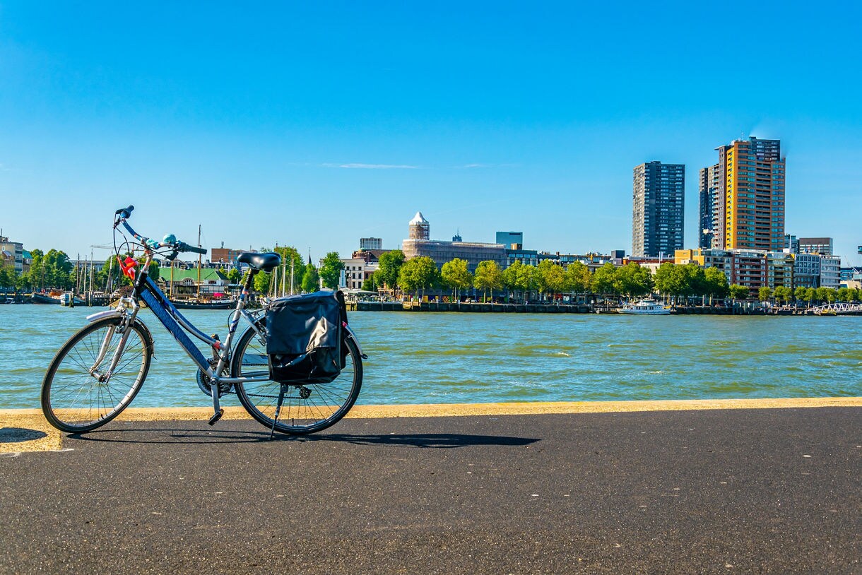  A bicycle parked along the Rotterdam waterfront with the Maas River in the foreground and modern high-rises rising behind leafy trees under a clear blue sky.