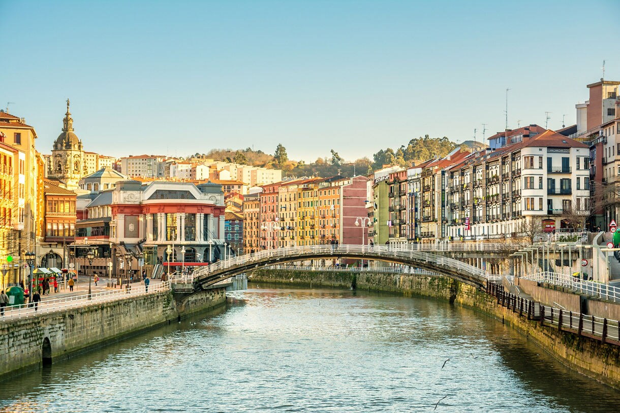 View of a pedestrian bridge crossing the Nervión River in Bilbao, lined with colorful historic buildings and a market hall under a clear sky.