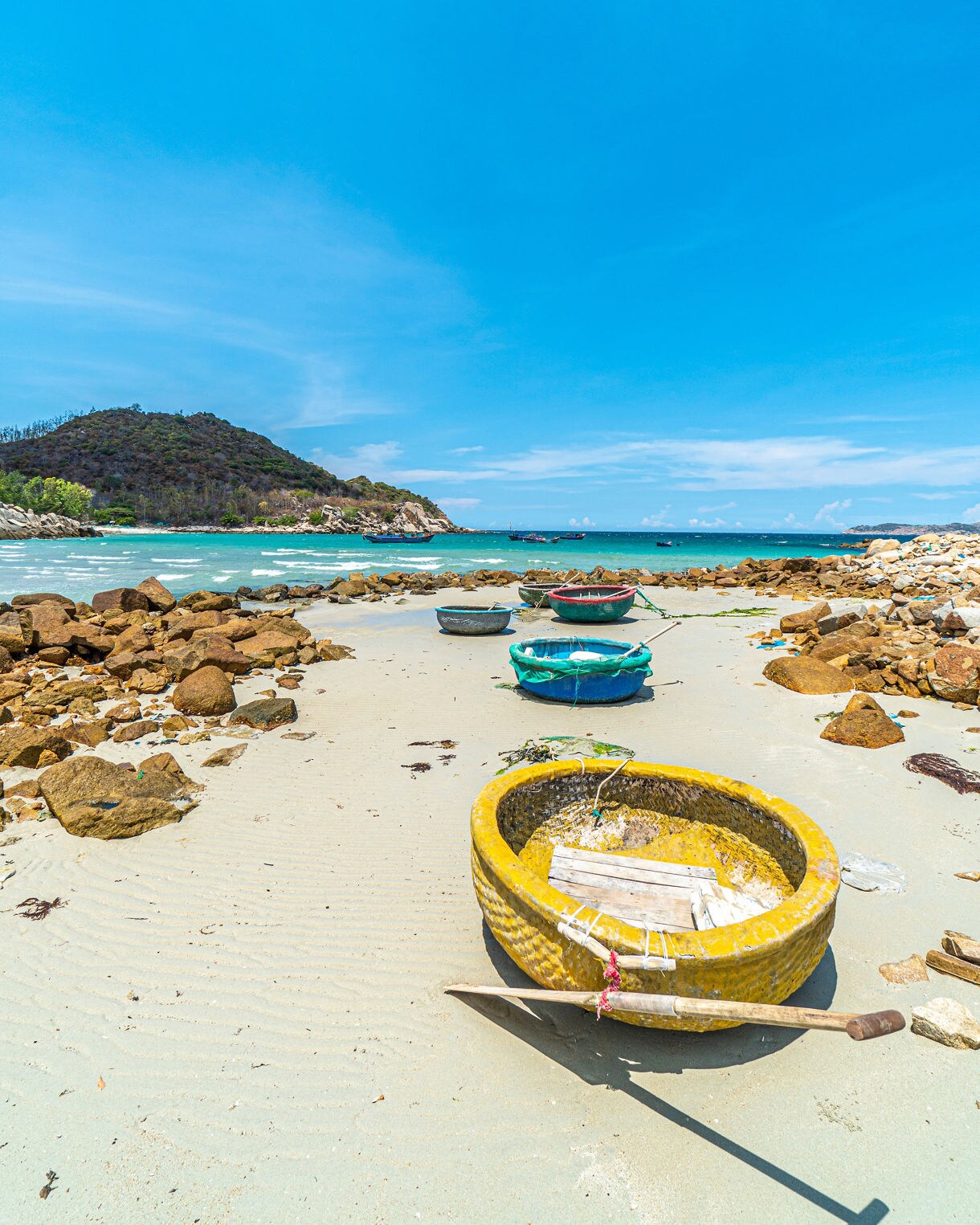 Round fishing boats in bright colors line the sandy shore of Binh Tien Beach, with turquoise water and rocky hills in the distance.