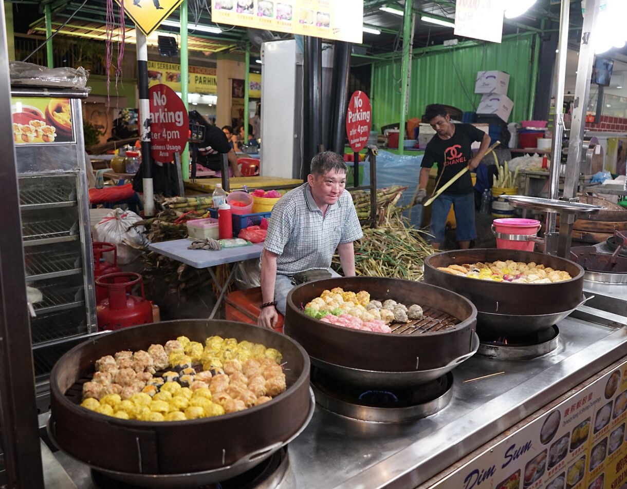 A street food vendor in Kuala Lumpur cooking dim sum in large steaming pans at a bustling night market stall.