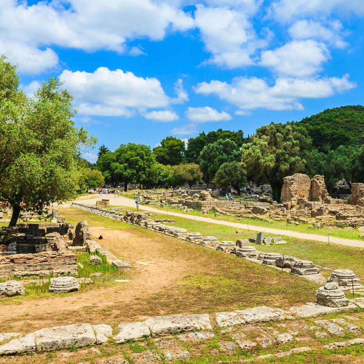 Ancient ruins surrounded by green trees and grassy paths at Olympia, Greece, under a bright blue sky with scattered clouds.