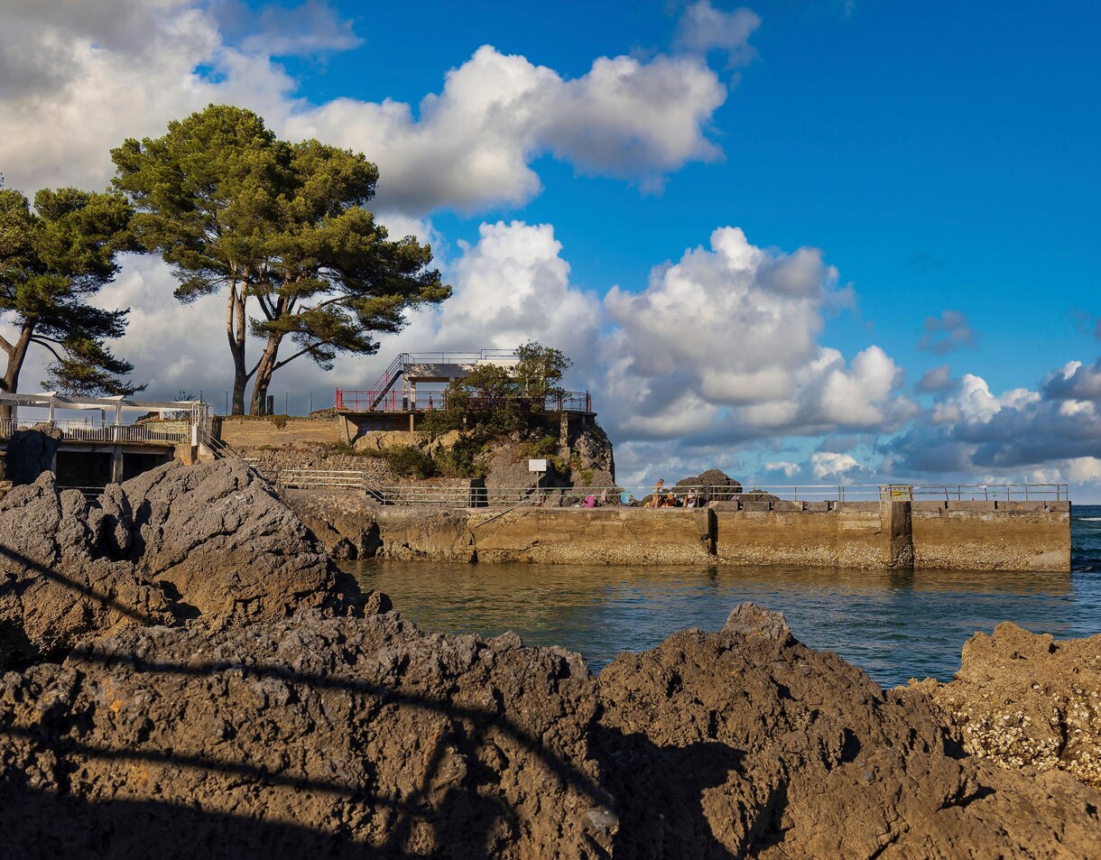 Coastal view along the Bay of Biscay featuring rocky cliffs, pine trees and a small pier under a bright blue sky with scattered white clouds.