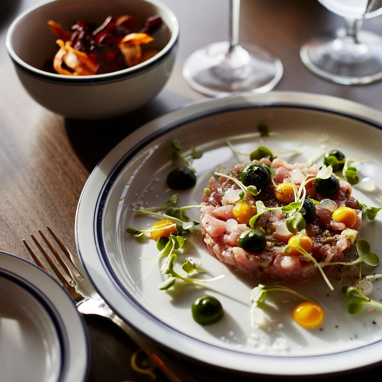 A gourmet starter with thinly sliced tuna adorned with microgreens, petite yellow tomatoes, and a drizzle of vibrant green sauce, accompanied by a side dish of red-leaf salad, all elegantly plated for fine dining.