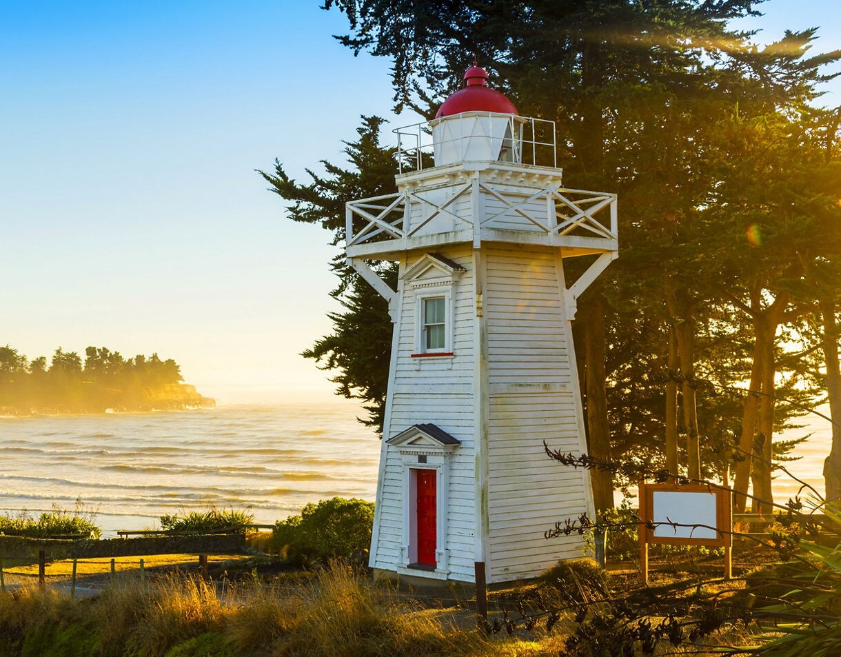 White wooden lighthouse with a red top overlooking rolling waves at sunset, surrounded by trees and golden coastal grasses.