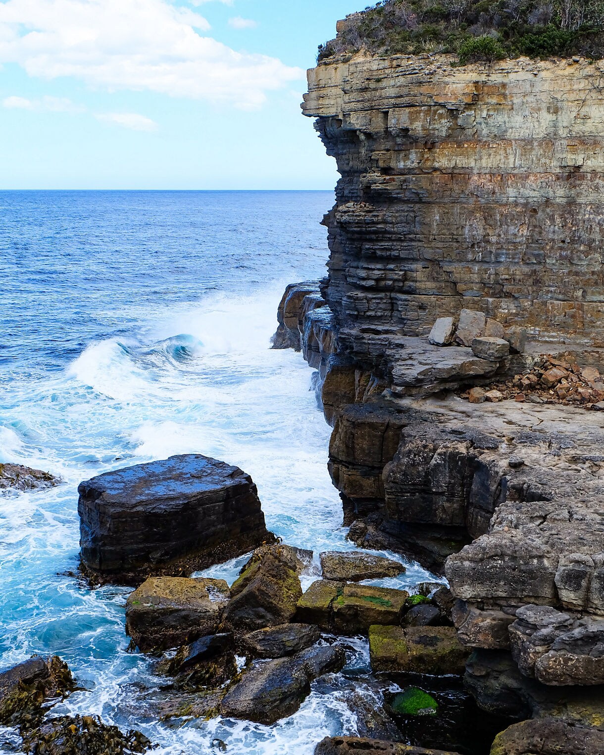 Rocky coastal cliffs with layers of stone overlooking turquoise waves crashing against boulders below.