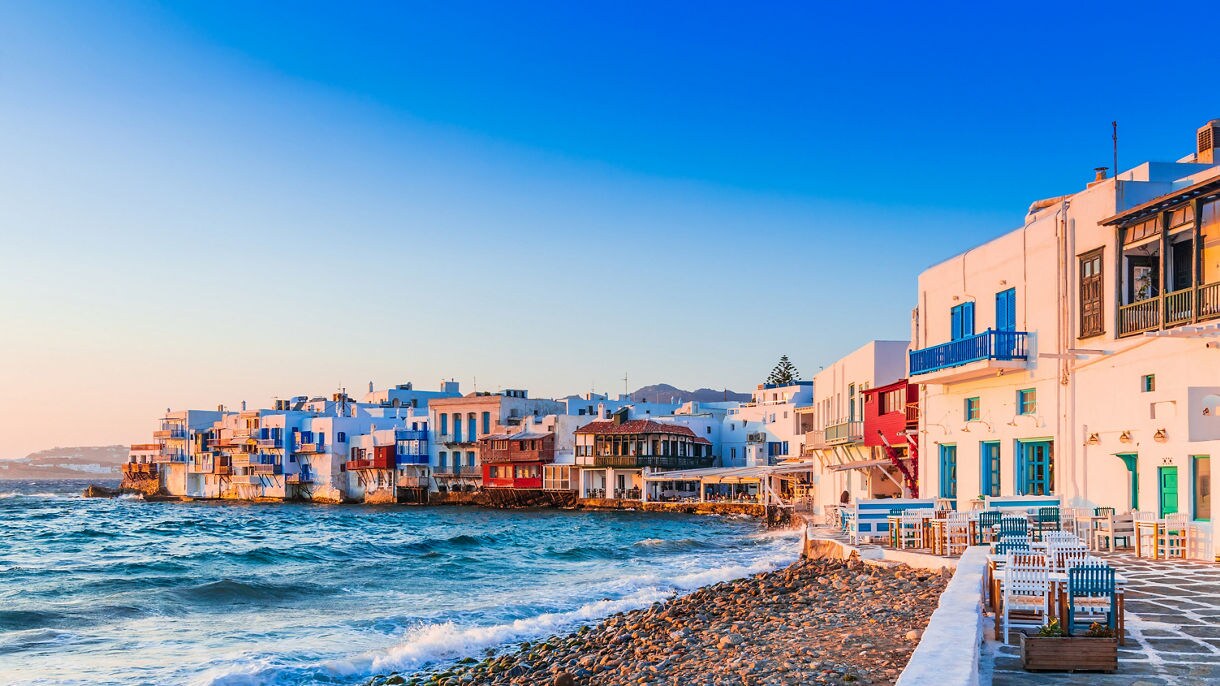 Whitewashed waterfront buildings with colorful balconies line a rocky shoreline in Mykonos, Greece, as waves roll in under a clear blue sky during golden hour.
