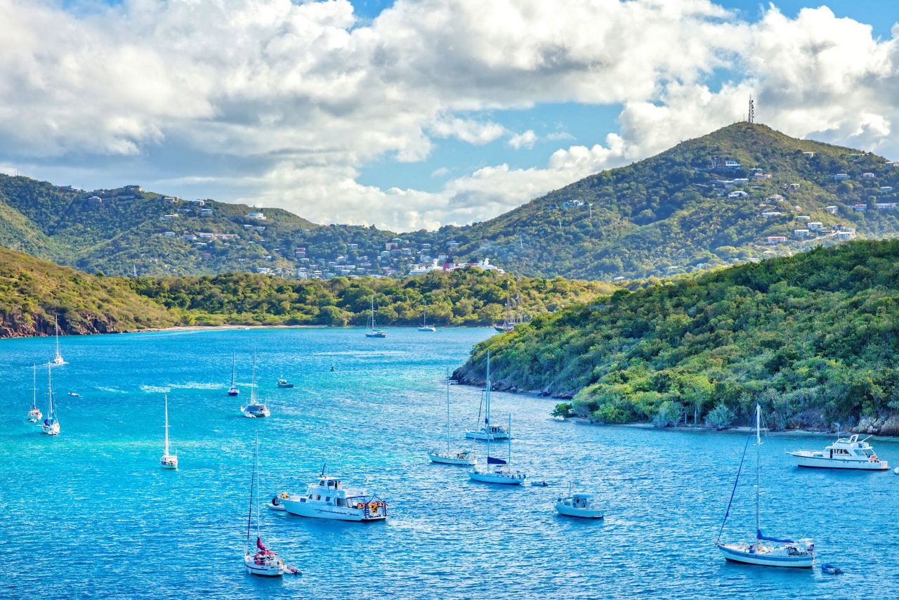 Multiple sailboats and yachts anchored in bright blue Caribbean waters surrounded by verdant hillsides dotted with homes