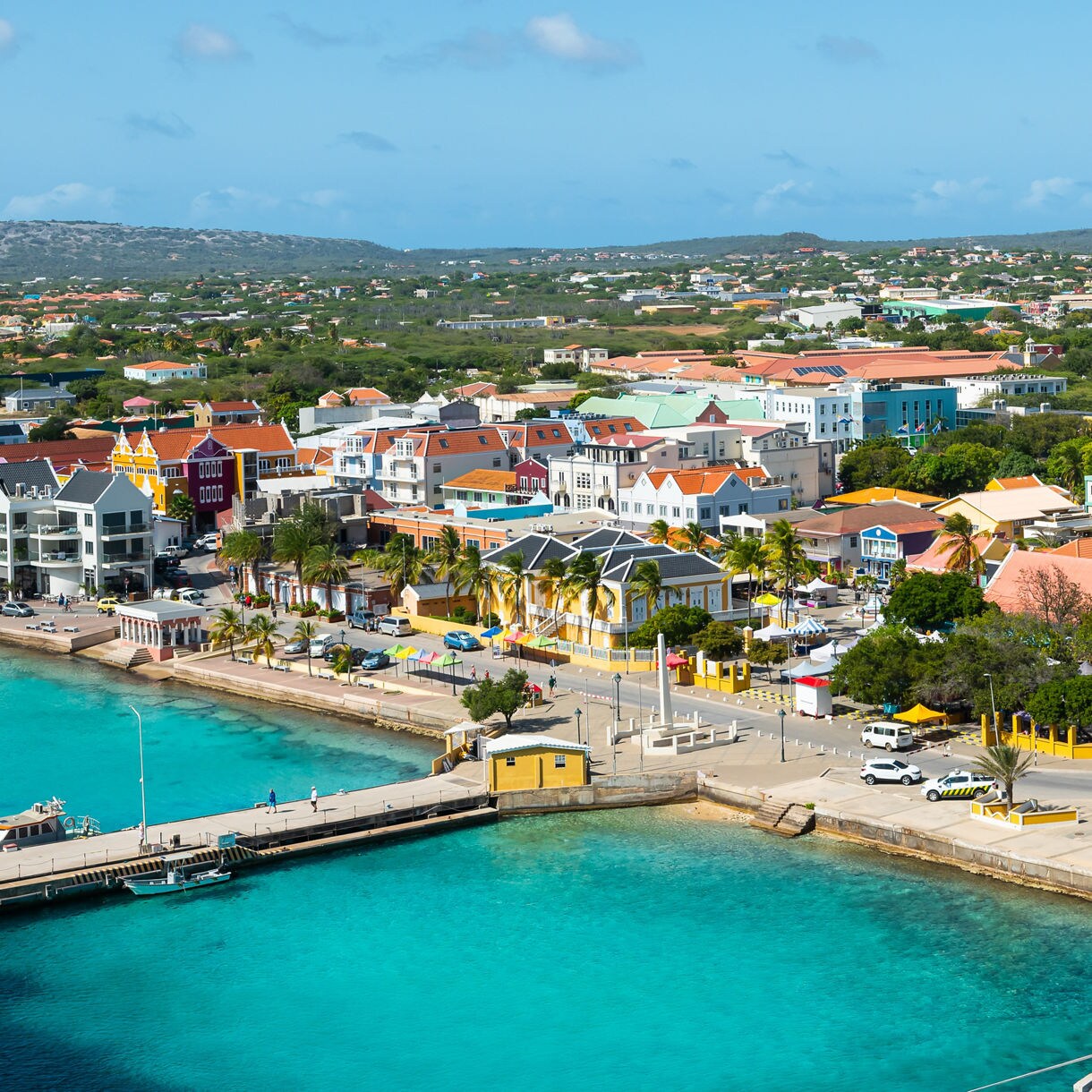  Aerial view of Bonaire’s colorful harbor town with pastel buildings lining turquoise water and low hills beyond.