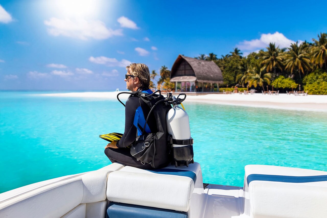 A diver with scuba gear sits on a boat, overlooking the pristine turquoise waters and a beach in the background, ready for a snorkel or dive experience in Bonaire