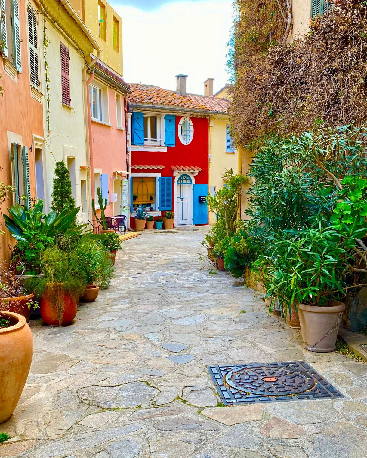 A charming stone alley in Bormes-les-Mimosas lined with colorful pastel houses, blue shutters and lush potted plants, leading to a bright red house at the end.