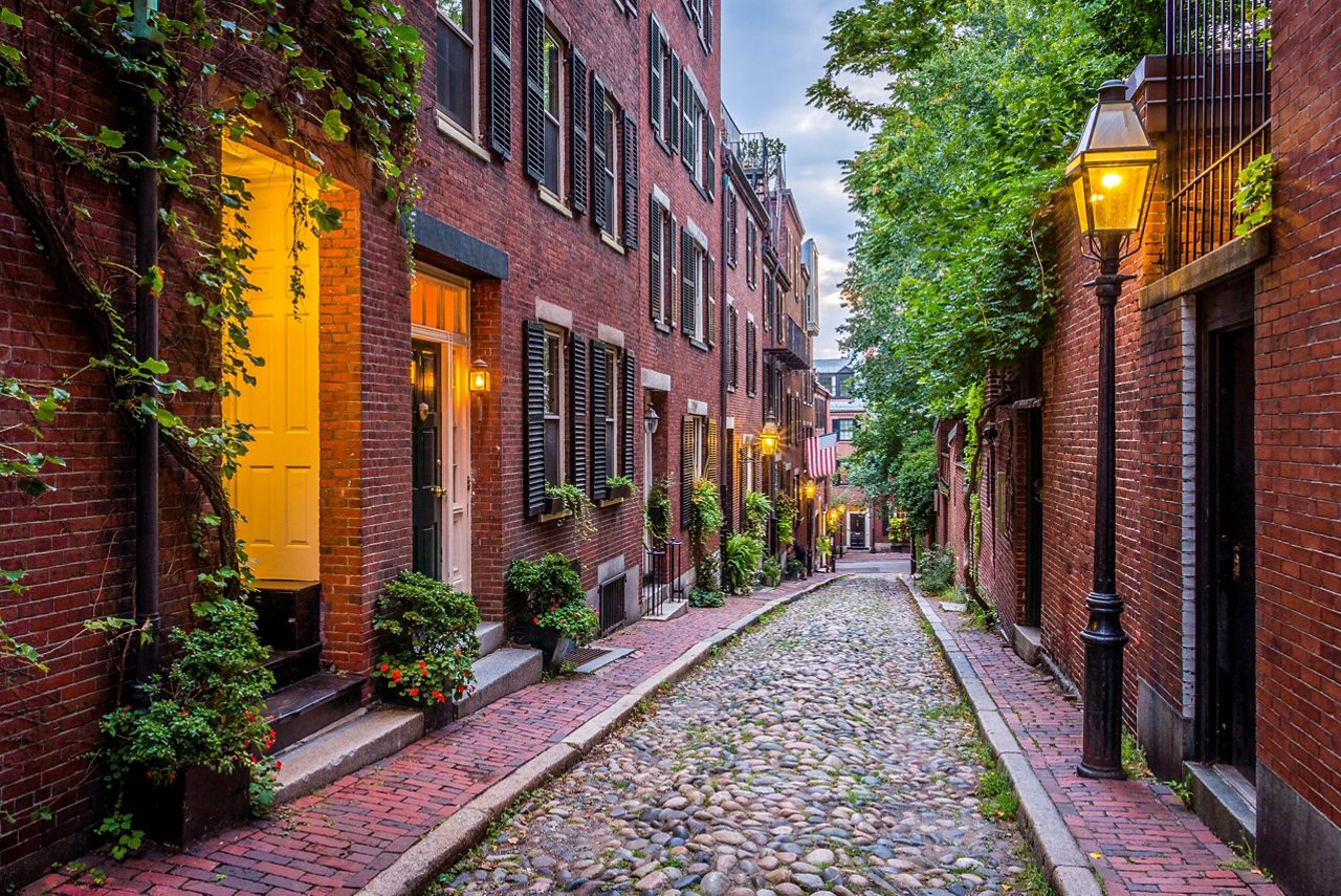 Imagine a picturesque historic street in Boston's Beacon Hill neighborhood, lined with gaslit lanterns, brick row houses, and ornate ironwork. The scene is set against a clear blue sky with wispy clouds, showcasing the area's well-preserved 19th-century architecture.