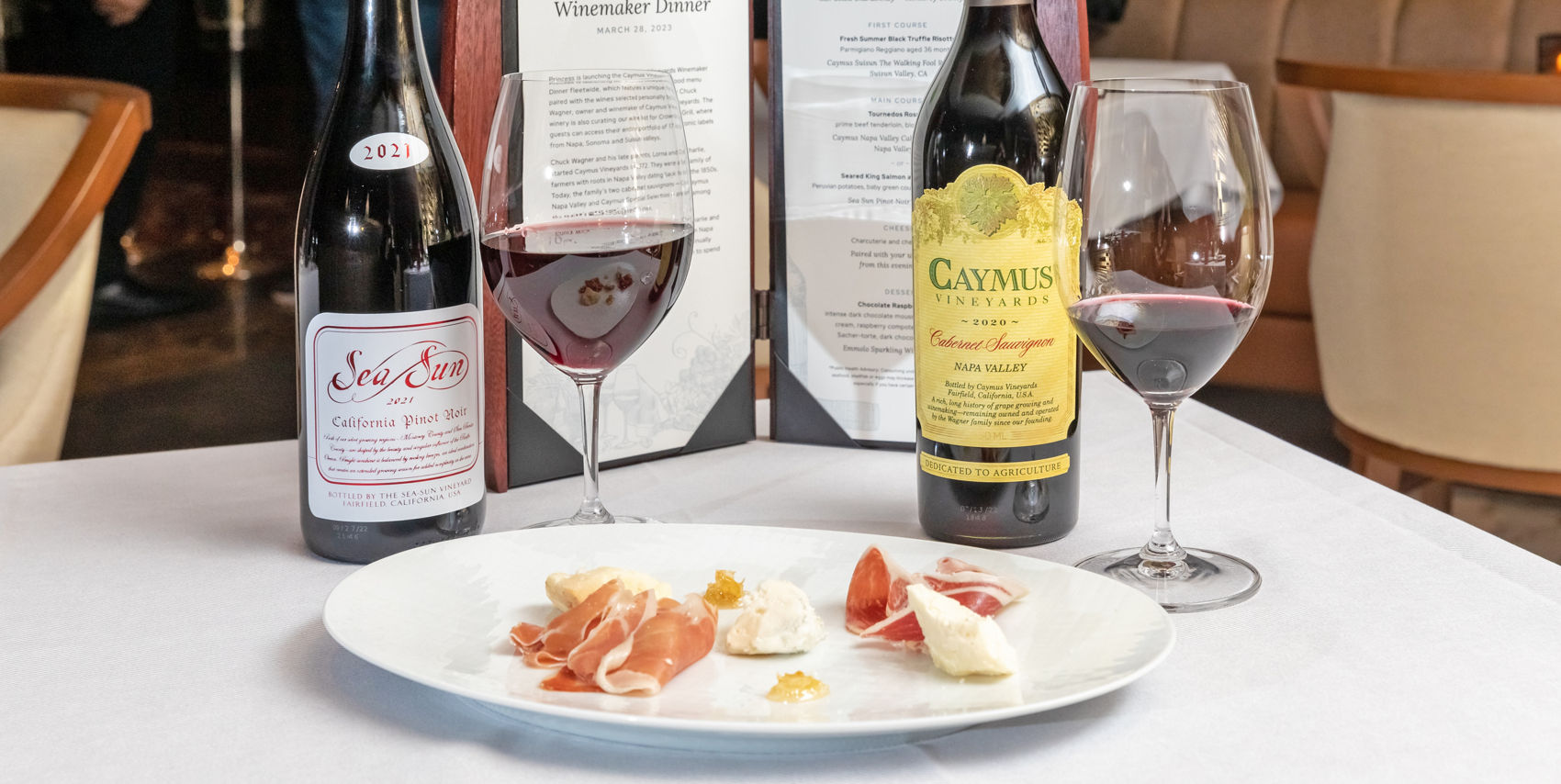 Two bottles of red wine — Sea Sun Pinot Noir and Caymus Cabernet Sauvignon — beside a glass of wine and a charcuterie plate on a white tablecloth.
