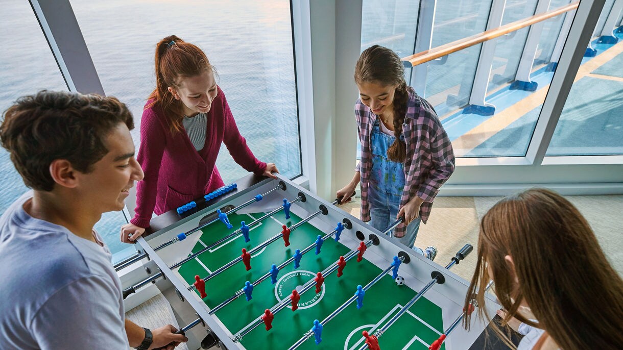 Four teenagers play a game of foosball together in a room with large windows overlooking the ocean.