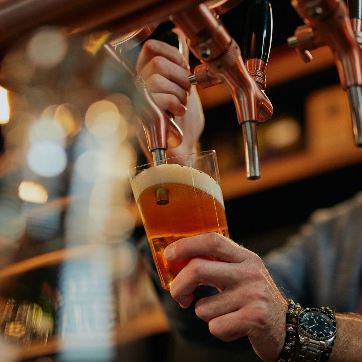 Close-up of a bartender filling a pint glass with beer from a tap, with golden foam forming at the top.