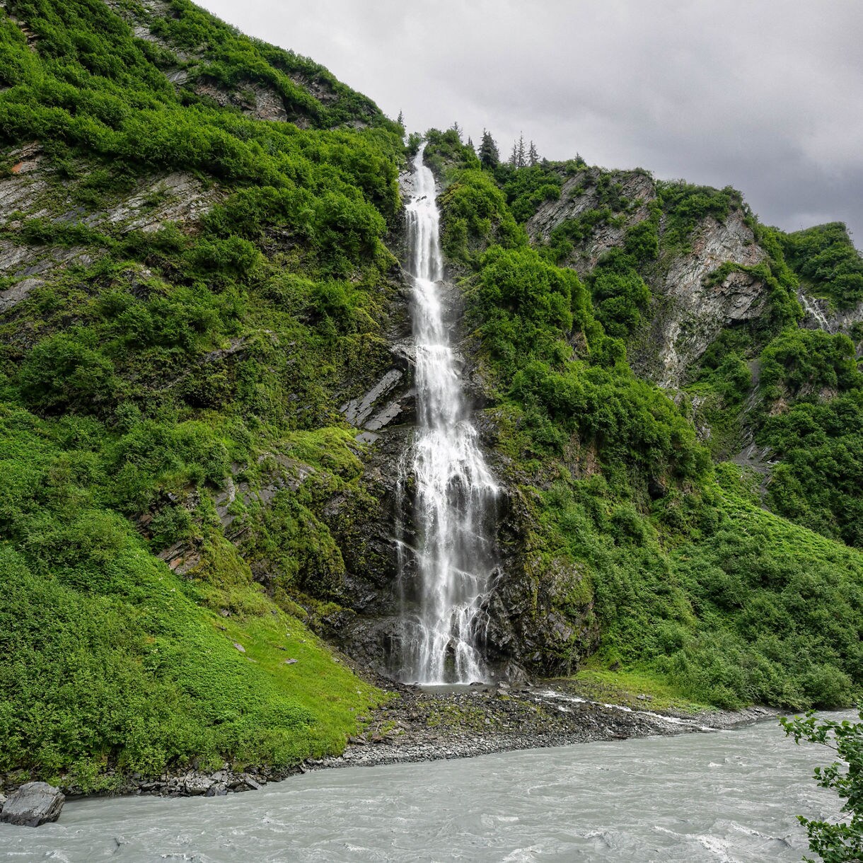 A serene waterfall cascades down a rocky cliff, surrounded by lush green vegetation and a flowing river below under a cloudy sky.