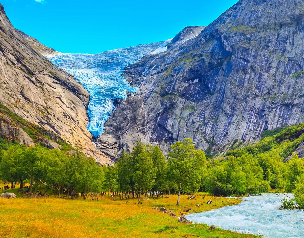 Briksdal Glacier in Norway, with a bright blue tongue of ice descending between steep rocky cliffs, overlooking a green meadow, forest and glacial river.