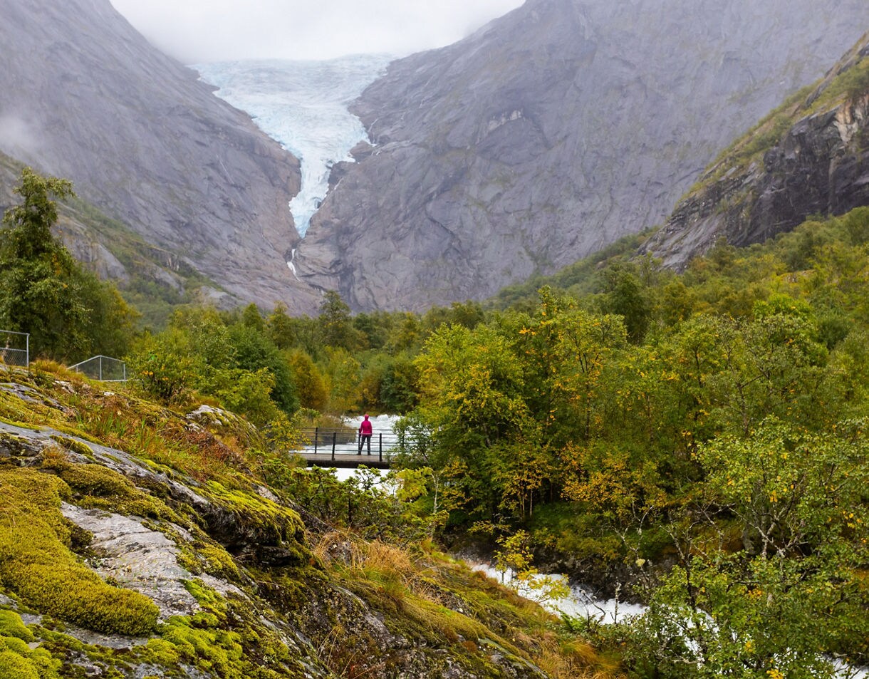 A hiker on a bridge near Briksdal Glacier in Norway, surrounded by mossy rocks, dense forest and towering cliffs with the glacier flowing between them.
