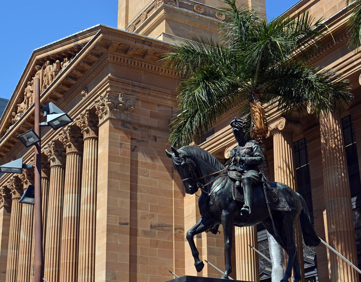 Brisbane City Hall’s sandstone columns and detailed pediment with an equestrian statue in the foreground, framed by palm trees under a clear blue sky.