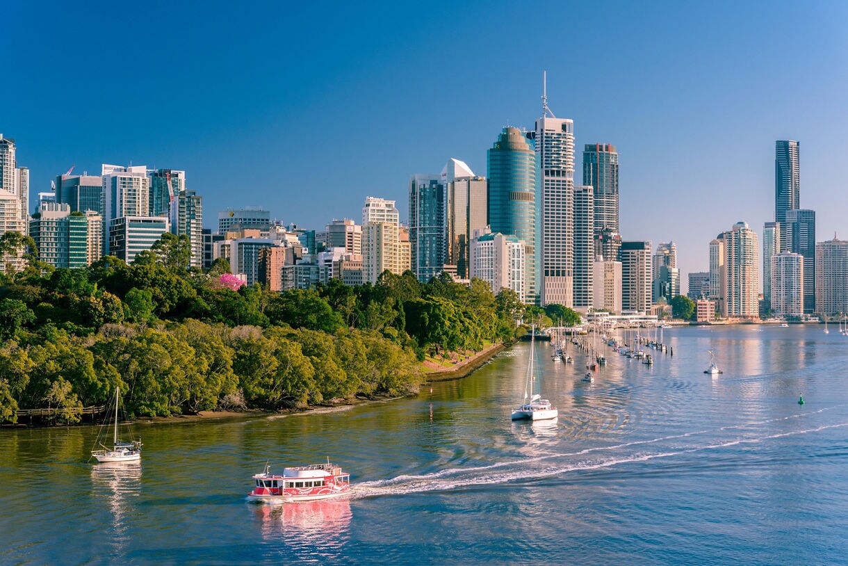 Brisbane skyline with modern skyscrapers, green parkland, and boats traveling along the Brisbane River.